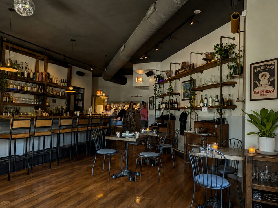 Warm, industrial-style wine bar interior with wood floors, high-top bar seating, wall shelves lined with bottles and glassware, and a few guests gathered near the back.